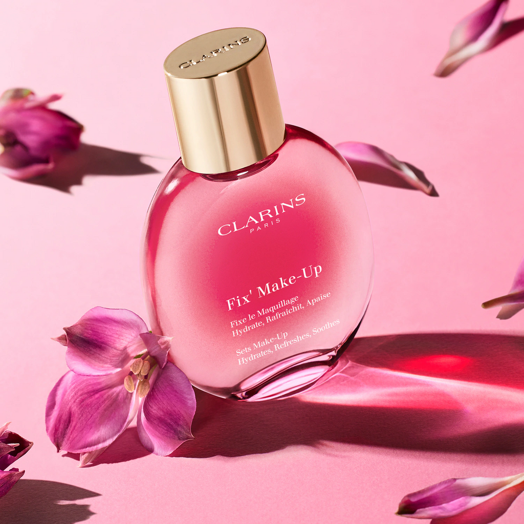 Clarins Fix’ Make-Up 50 ml entouré de fleurs roses sur un fond rose ensoleillé.