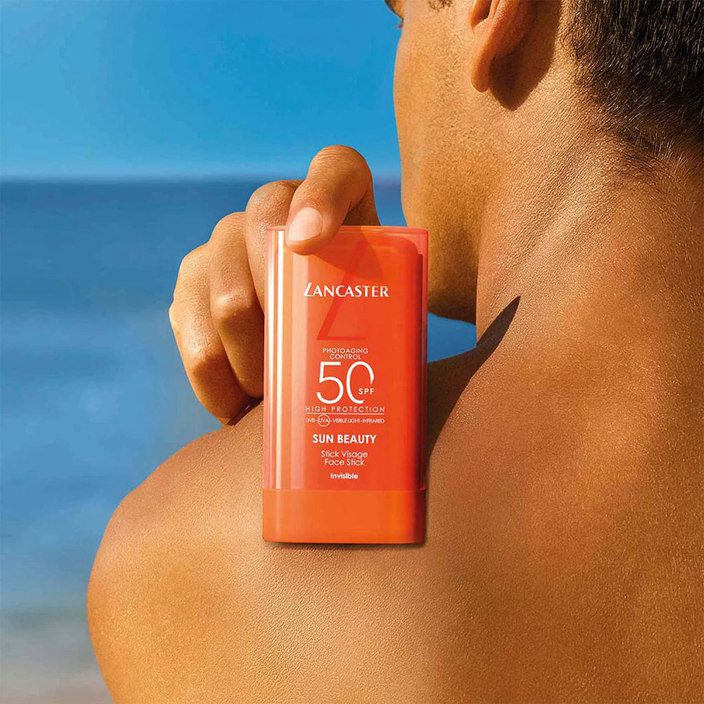 Personne tenant le stick Lancaster Sun Beauty SPF 50 contre son épaule, avec la mer en arrière-plan